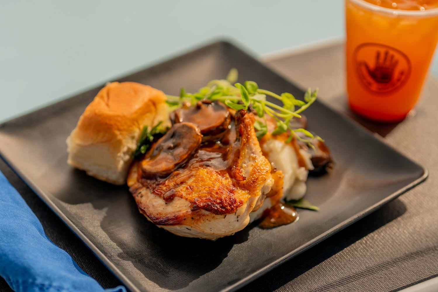 Grilled chicken with mushrooms, roll, and greens on black plate, drink in background.