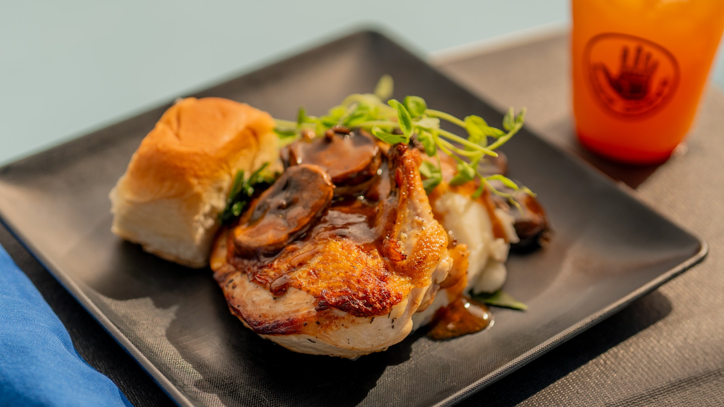 Grilled chicken with mushrooms, roll, and greens on black plate, drink in background.