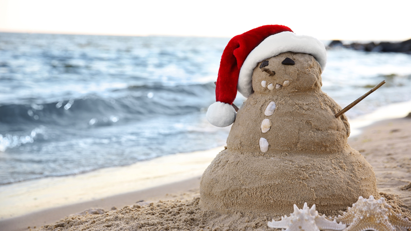 Sand snowman with Santa hat on beach by the ocean.
