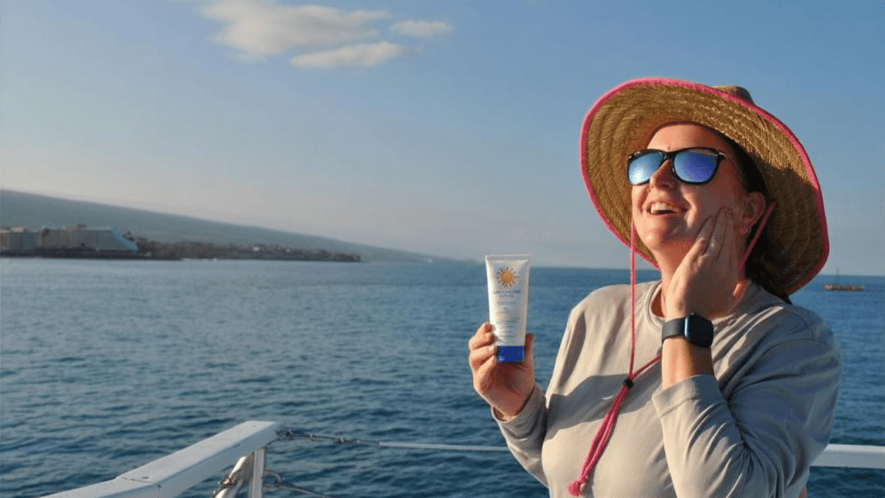 Person in a hat smiling, standing on a boat applying reef safe sunscreen near the ocean.