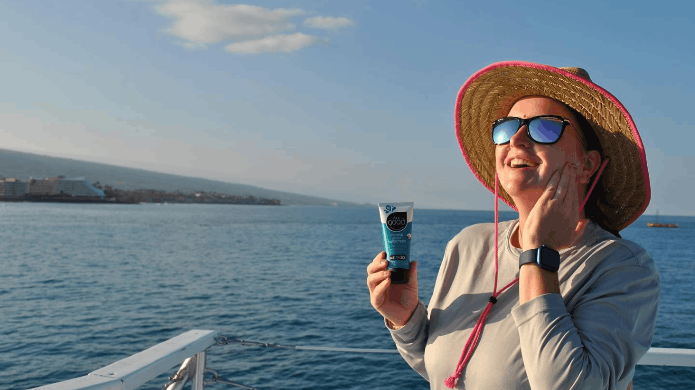Person in a hat smiling, standing on a big island eco-friendly tour applying reef safe sunscreen near the ocean.