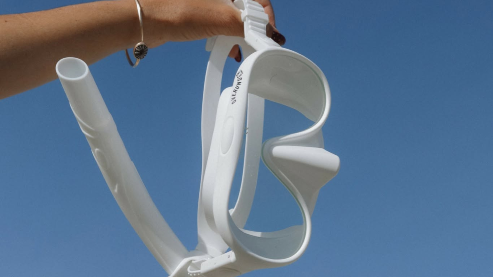 Person holding white snorkel mask against a clear blue sky.