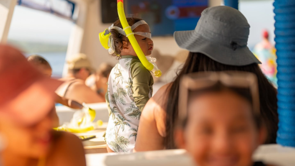 Child with snorkel gear on a boat with adults in hats, blurred background.